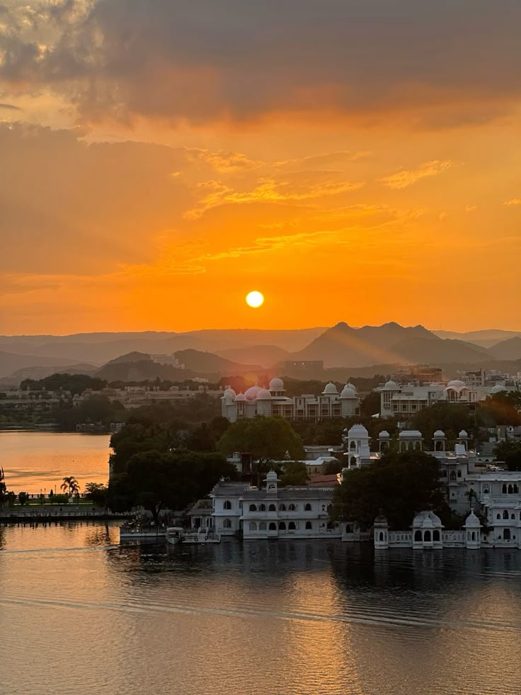 Sunset over a city with buildings reflected in water, mountains in the background